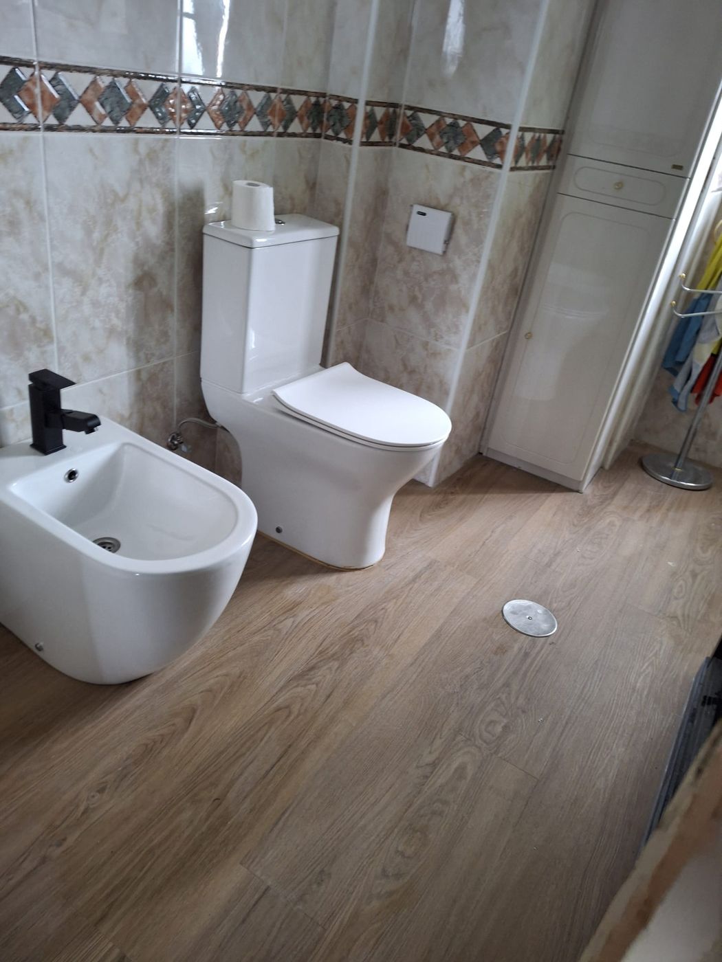 Baño con inodoro y bidé en blanco, suelo imitación madera y alicatado en tonos beige