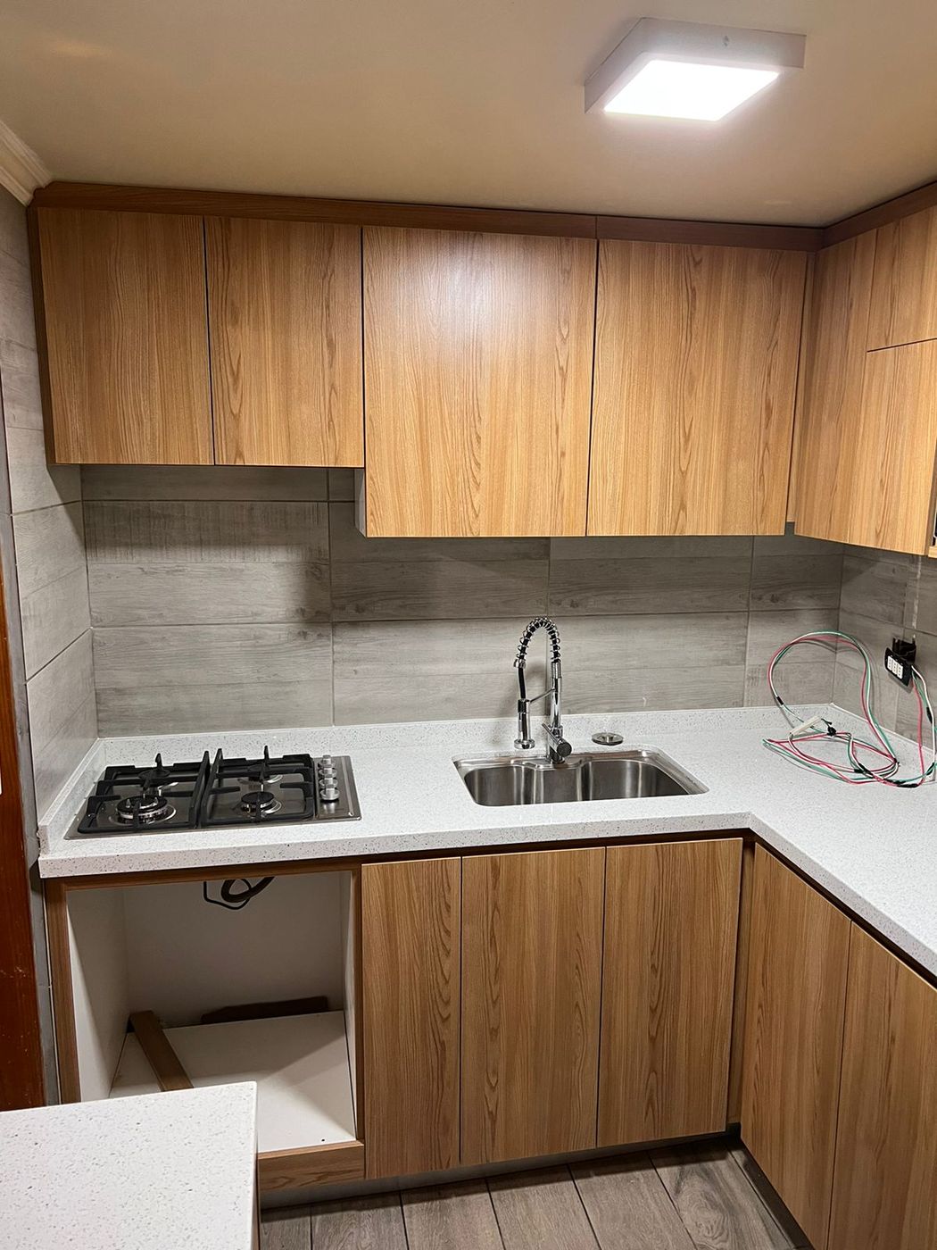 Cocina en L con muebles de madera clara, placa de gas de dos fuegos y fregadero inoxidable